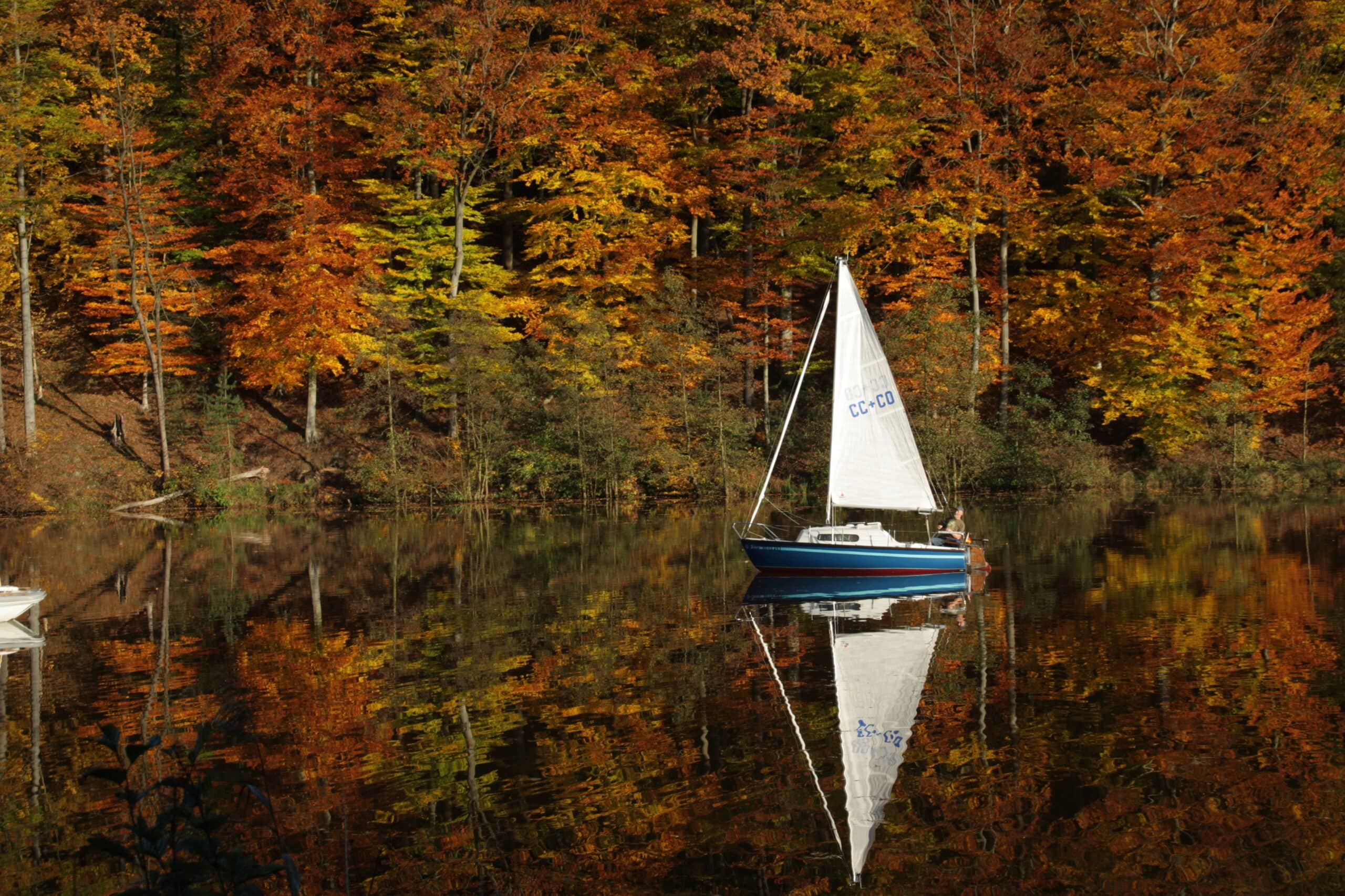 Un voilier et une forêt automnale aux couleurs rouges, jaunes et vertes se reflètent sur la surface du lac.